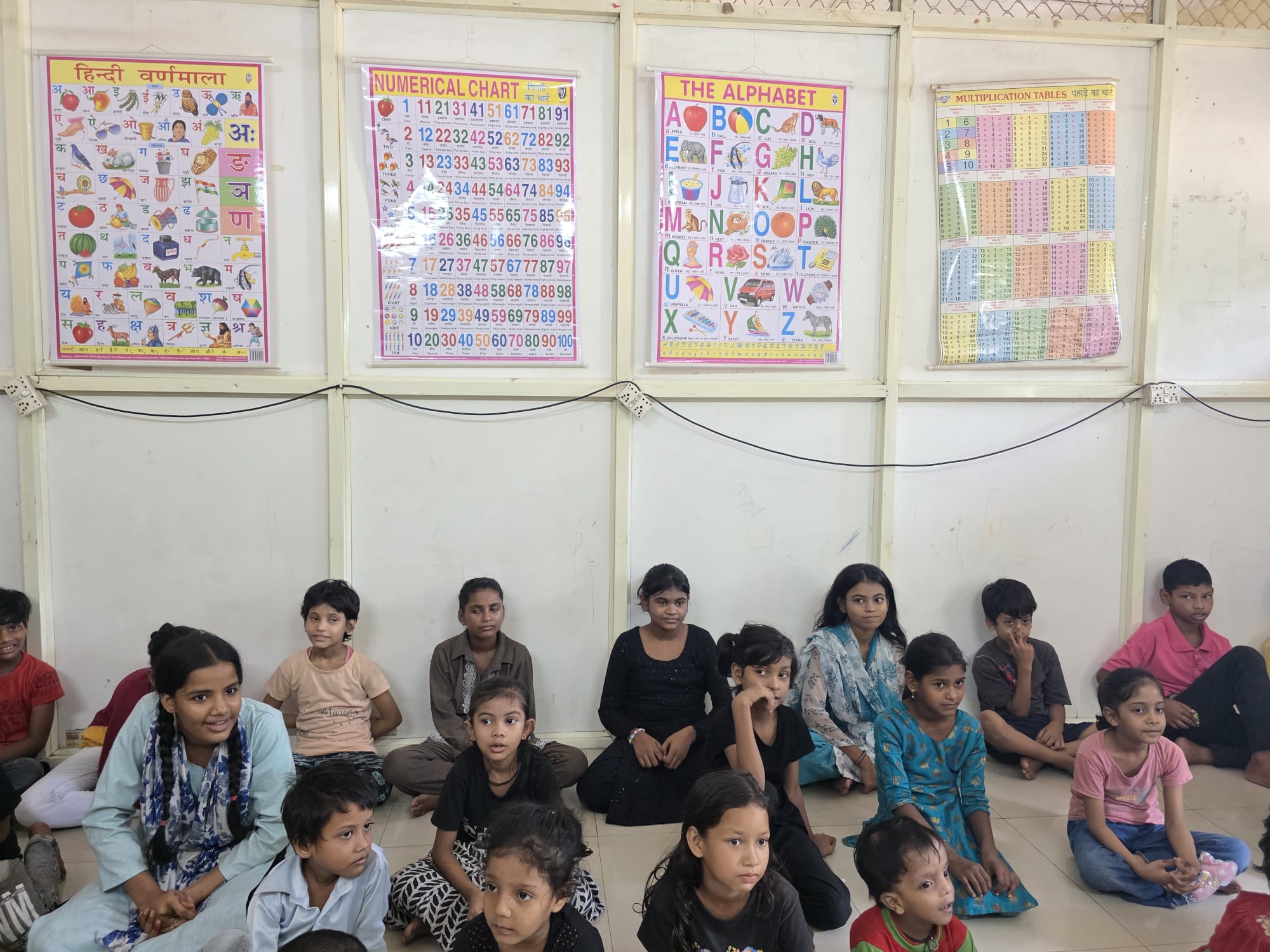 Children seated in a classroom below Hindi and English learning charts