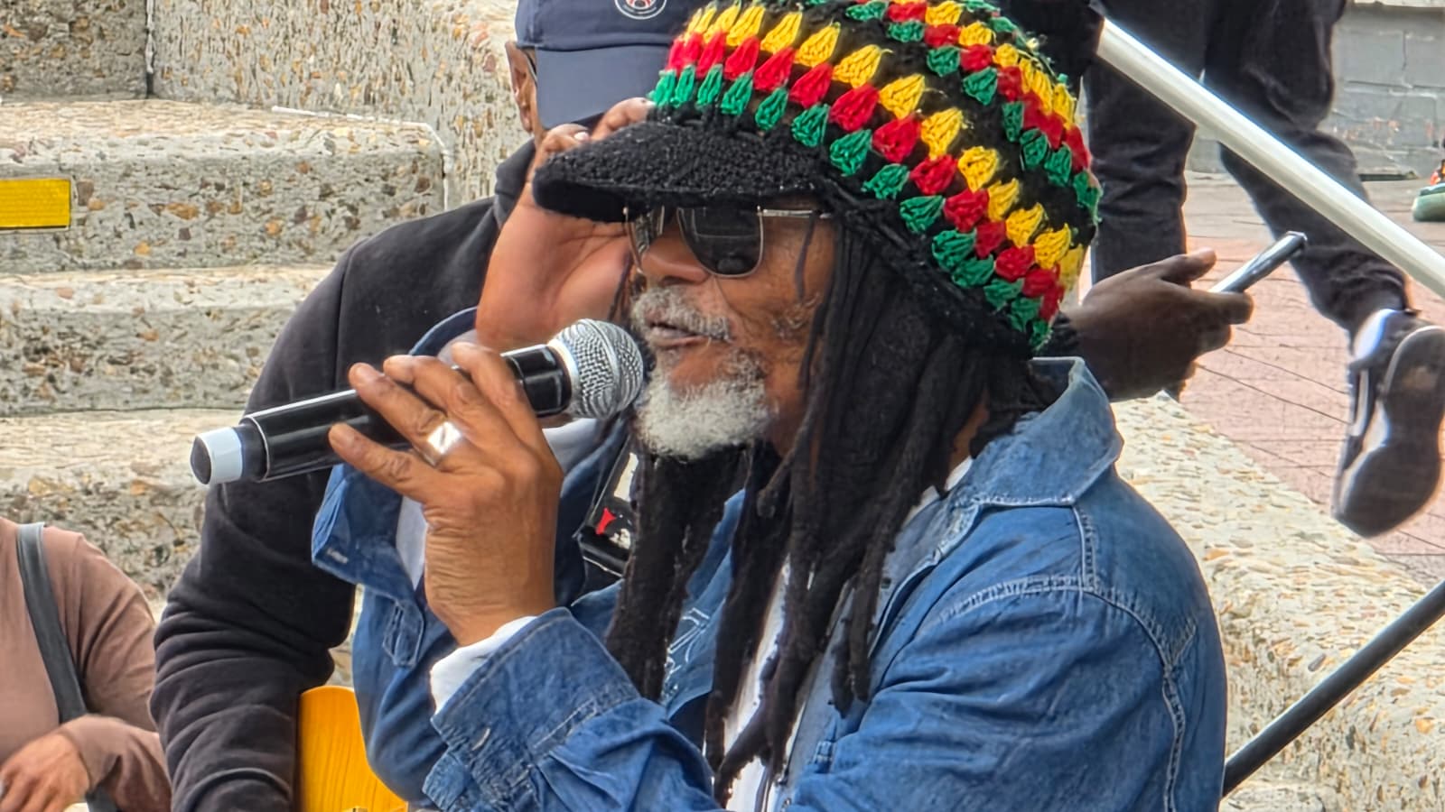 A community member in a knitted rasta hat speaking into a microphone at an outdoor gathering in Cape Town